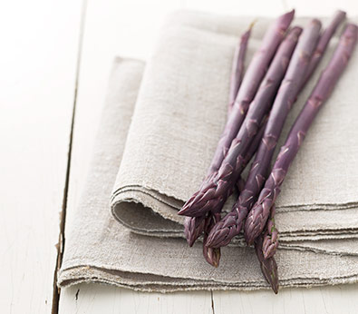 6 Purple Passion asparagus shoots resting on some beige linen napkins atop white wood
